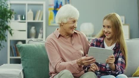 cheerful andsmiled grandmother and granddaughter sitting together on the sofa at home and watching something on the tablet device