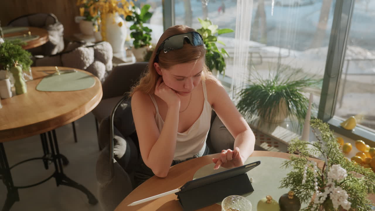 Hvid kvinde bruger tablet på solbeskinnet café, smilende mens hun tjekker beskeder og trykker på skærmen, latte og blomsterarrangement på rundt bord, solbriller på hovedet, afslappet stemning med varmt vindueslys