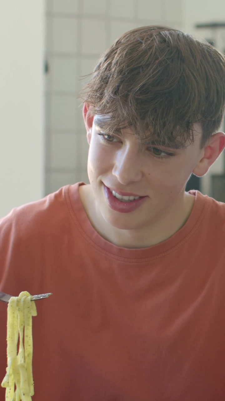 Video verticale: Si vede una forchetta che solleva gli spaghetti, uomo sorridente che si china per mangiare in cucina, maglietta arancione