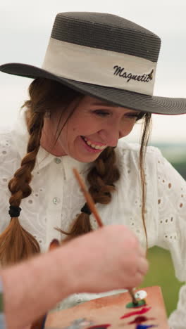 un artista, sosteniendo una paleta de pintura, trabajando en un gran lienzo en un entorno al aire libre. a su lado, una mujer con un vestido blanco y sombrero está de pie, sonriendo alegremente mientras observa su trabajo