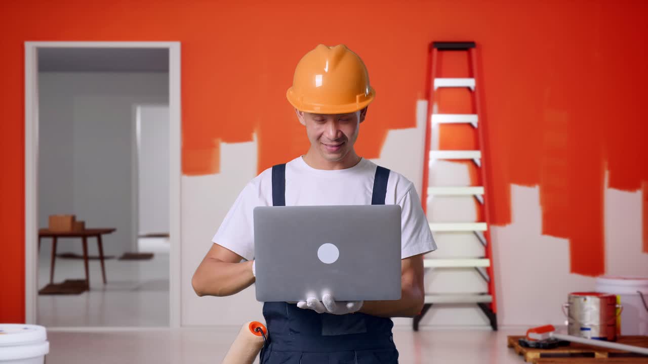 Uomo asiatico pittore con casco di sicurezza che usa un laptop e sorride, in piedi con un muro in fase di pittura nella stanza
