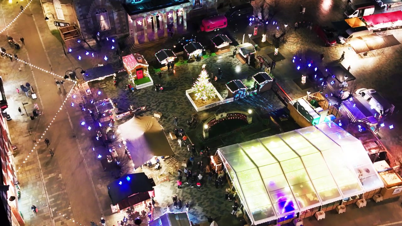 Luftoptagelser med drone om natten af Salisbury torv med oplyste julemarkedsboder, et juletræ og det historiske Guildhall oplyst.