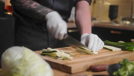 fresh zucchini being sliced on a wooden board by young professional male chef in an elegant black shirt with tattoos