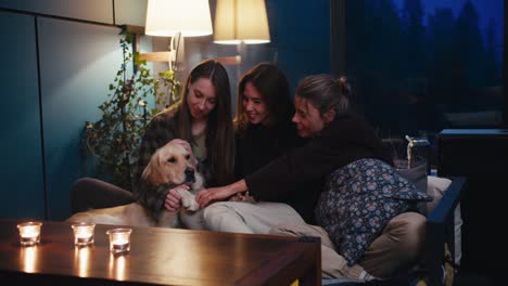a trio of girls are sitting on a dewan and chatting, they are playing with their light-colored dog in a cozy room in the evening