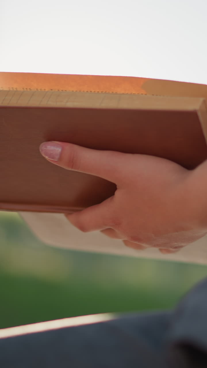 빛은 배경의 흐릿한 초록색에서 빛나고, 밖에서 앉아있는 여성이 들고 있는 책에서 반사되며, 그녀는 검은색 에 안경을 착용하면서 페이지를 부드럽게 뒤집습니다.