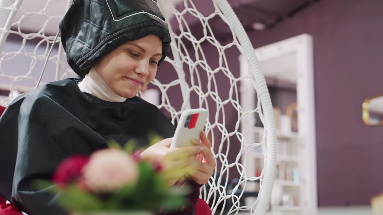 미용실에서 헤어 드라이어 아래에 앉아 휴대폰을 사용하는 여성, 케이프를 두르고 조용한 기다림을 즐기는 모습, 차분한 색감과 꽃무늬 배경, 스타일리스트가 다른 손님을 보는 동안 메시지를 확인하며 스크롤하는 모습