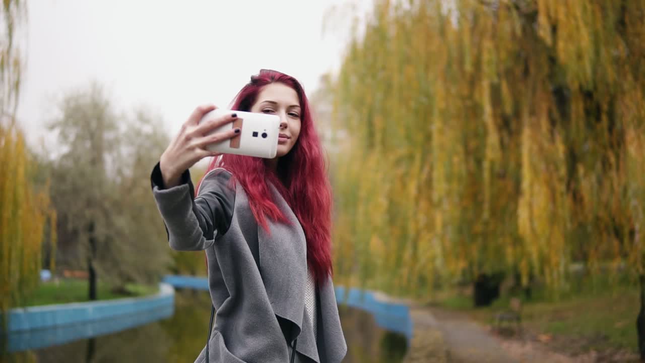 joven atractiva con sombrero rojo haciendo una selfie en su teléfono inteligente mientras estaba de pie junto a un estanque artificial en un parque de otoño