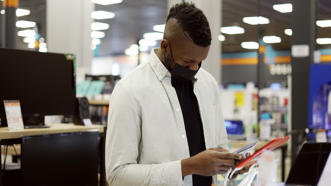 African American man in mask is choosing a new mobile phone in a shot, checking how it works
