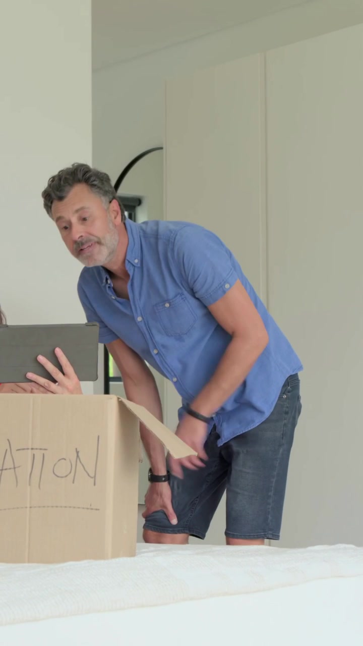Video vertical: Hombre empacando con camisa azul y pantalones cortos llenando una caja de DONACIÓN en la cama, revisando una tableta