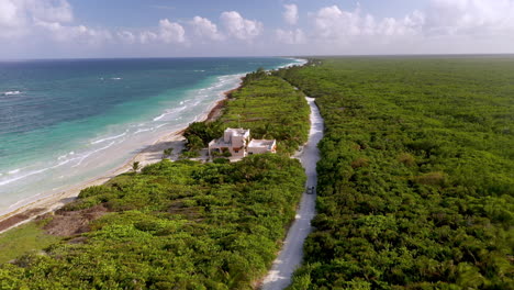 Drone-shot-of-car-driving-along-the-coastline-near-Mahahual-Mexico-with-single-home,-dense-forest-and-clear-ocean-water