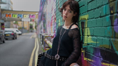 retrato de moda al aire libre de una mujer joven de estilo alternativo apoyándose en una pared cubierta de graffiti en la calle de la ciudad de londres, reino unido, en tiempo real.