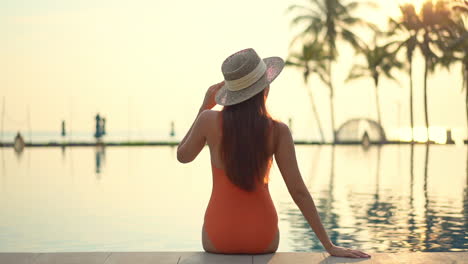 back view of fit female model wearing an orange monokini swimwear suit and hat sits on the edge of the swimming pool in turkey at sunset