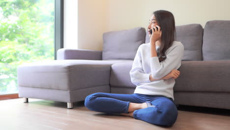 woman talking on phone, smiling asian thai girl sitting on floor next to sofa in casual clothes and having a flirting mobile phone conversation