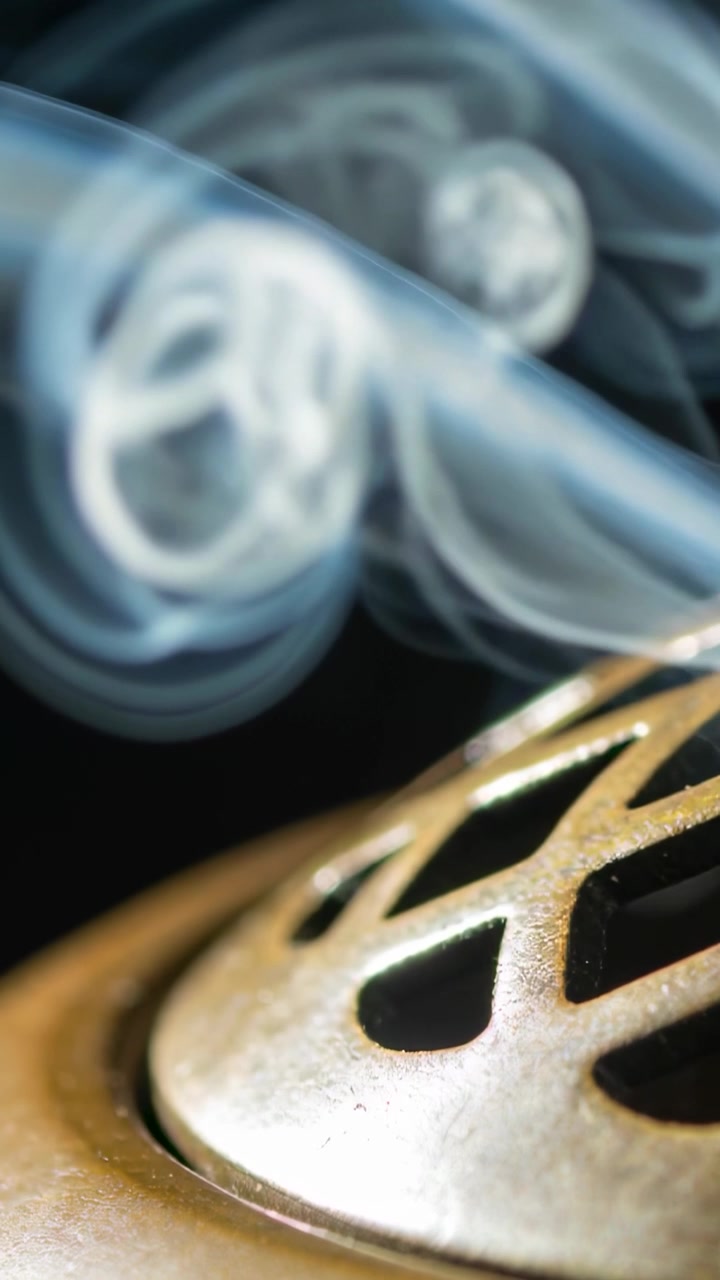 Vertical video: Incense burning inside metal vent releasing curling smoke on black backdrop