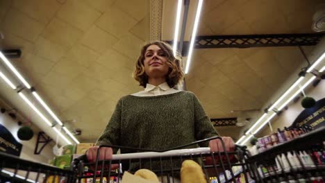 Pretty-woman-in-casual-clothes-is-walking-in-grocery-store-steering-shopping-trolley-with-bread-inside-it-and-looking-around-at
