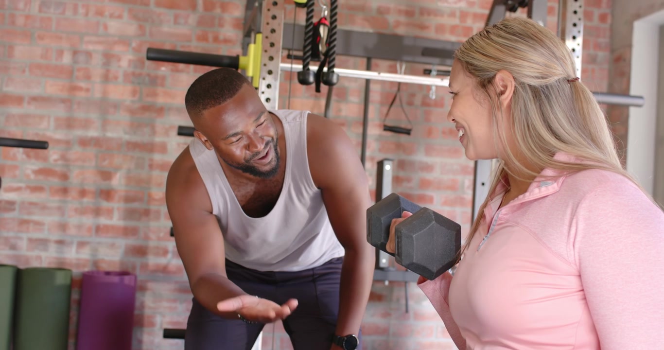 Ein männlicher Trainer beugt sich vor und leitet eine sportlich gekleidete weibliche Trainierende an, die im Fitnessstudio Bizepscurls mit einer sechseckigen Hantel ausführt, um die korrekte Form zu gewährleisten.