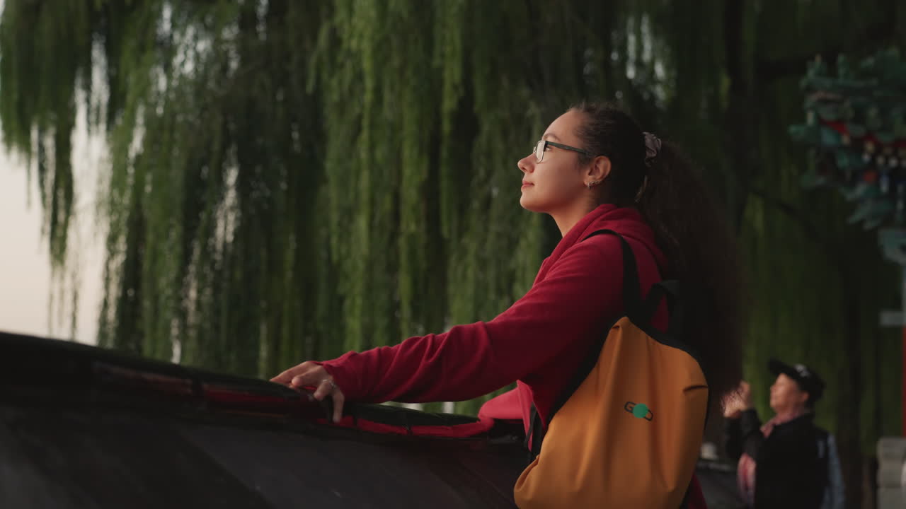 Giovane donna al crepuscolo accanto a un salice, appoggiata a una ringhiera, con zaino giallo e maglione rosso. Guarda il fiume in direzione di un padiglione, in una luce dorata e soffusa. Atmosfera tranquilla da parco urbano, una folla distante, atteggiamento contemplativo.