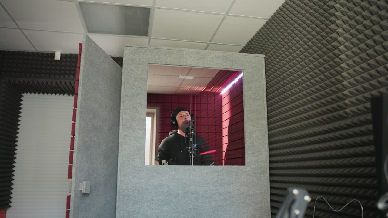 A man dressed in black stands in front of a studio microphone, wearing headphones as he sings passionately with hand movement in a red soundproofed room. The shot includes a speaker in the background