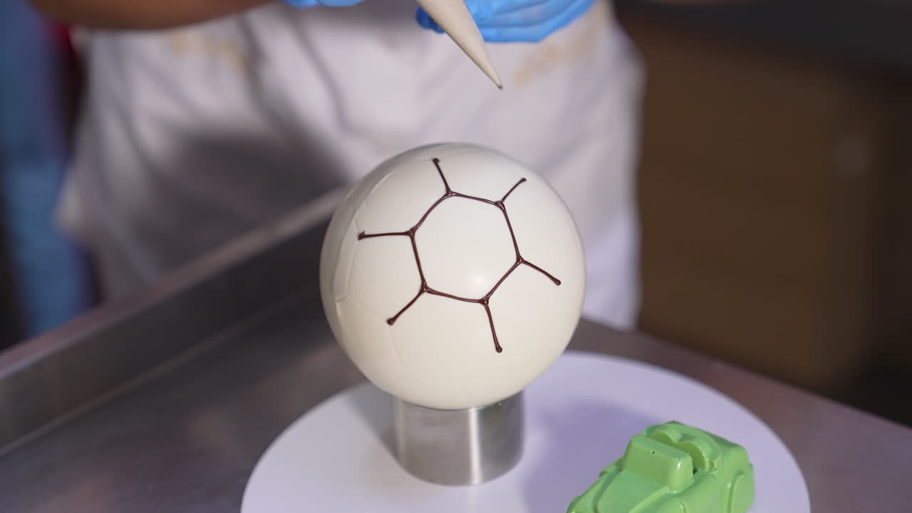 Confectioner decorates ball-shaped white cake with chocolate. Producing desserts at factory close up. Blurred backdrop.