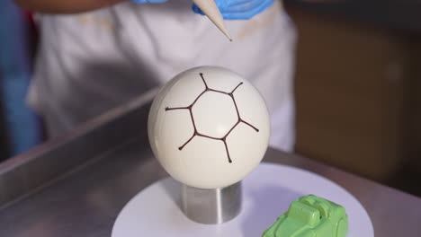confectioner decorates ball-shaped white cake with chocolate. producing desserts at factory close up. blurred backdrop.