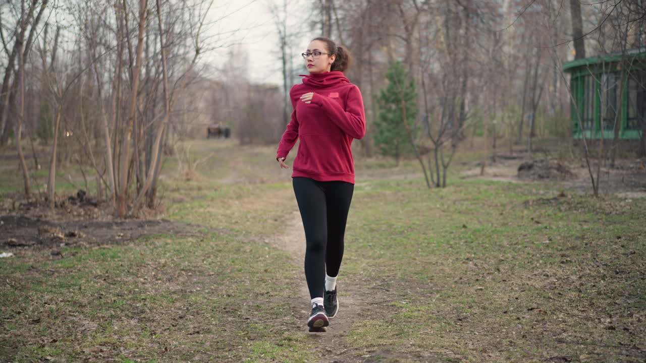 Morning Exercise Along Treelined Pathway, Woman Maintains Steady Pace During Suburban Trail Run, Focused Female Runner In Red Sweatshirt Moving Steadily Through Tranquil Wooded Suburban Trail