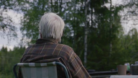 elderly person with white hair sitting alone outdoors on a striped chair facing lush green forest in quiet reflective moment