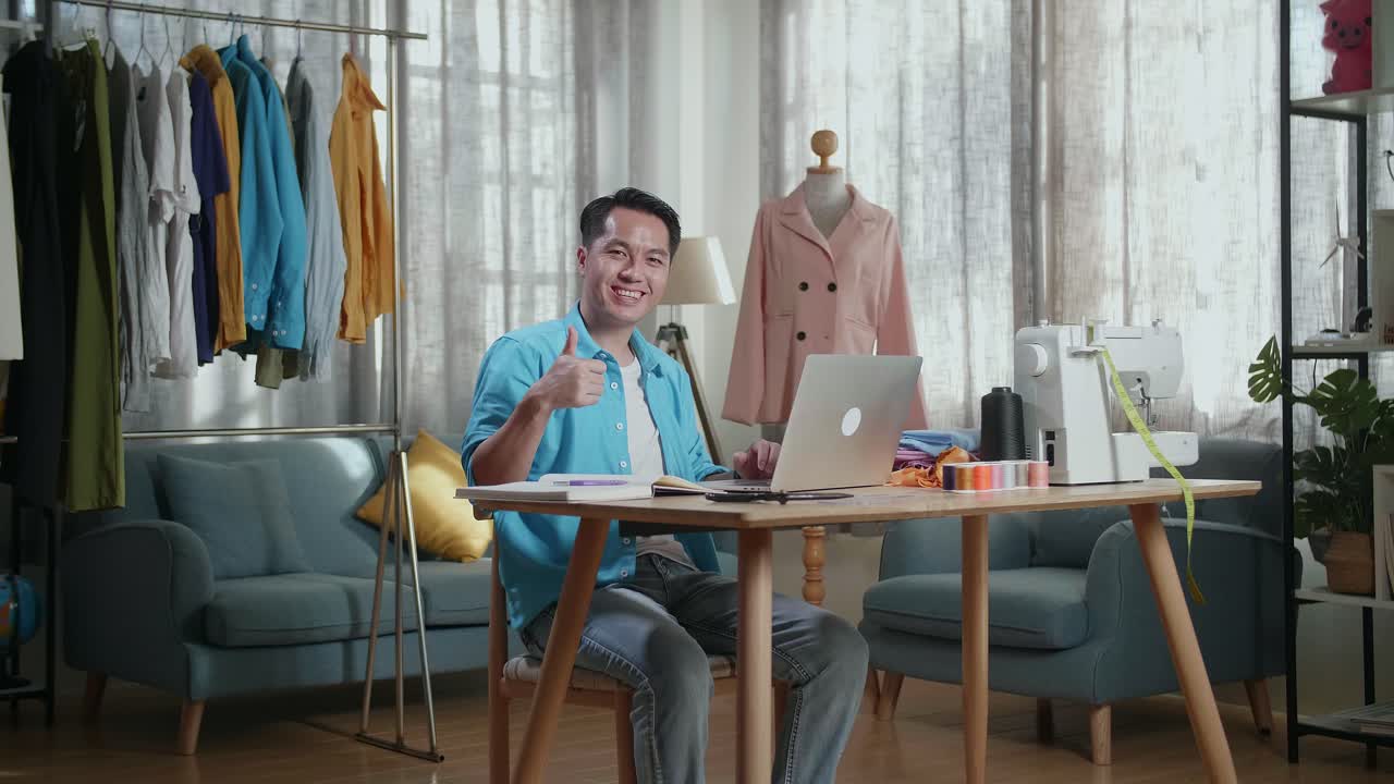 diseñador masculino asiático trabajando en una computadora portátil y sonriendo y levantando el pulgar hacia la cámara mientras diseña ropa en el estudio