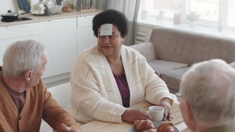 high angle of friends sitting at table, playing forehead detective game with sticky notes on head, african woman asking questions of her character, caucasian man answering, all smiling, applauding