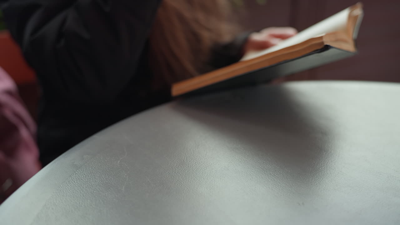 tazza di carta sul tavolo di un bar lettore, primo piano di una tazza di carta con coperchio e protezione testurizzata, mano che prende un caffè da asporto mentre sfoglia le pagine di un romanzo, luce mattutina soffusa, vapore delicato, atmosfera urbana rilassata
