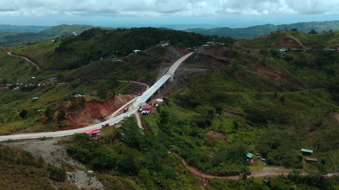 Drone footage showing an elevated bridge and mountain road under construction in the Philippines, with workers, machinery, and rugged terrain surrounded by green hills and dramatic cloudy skies