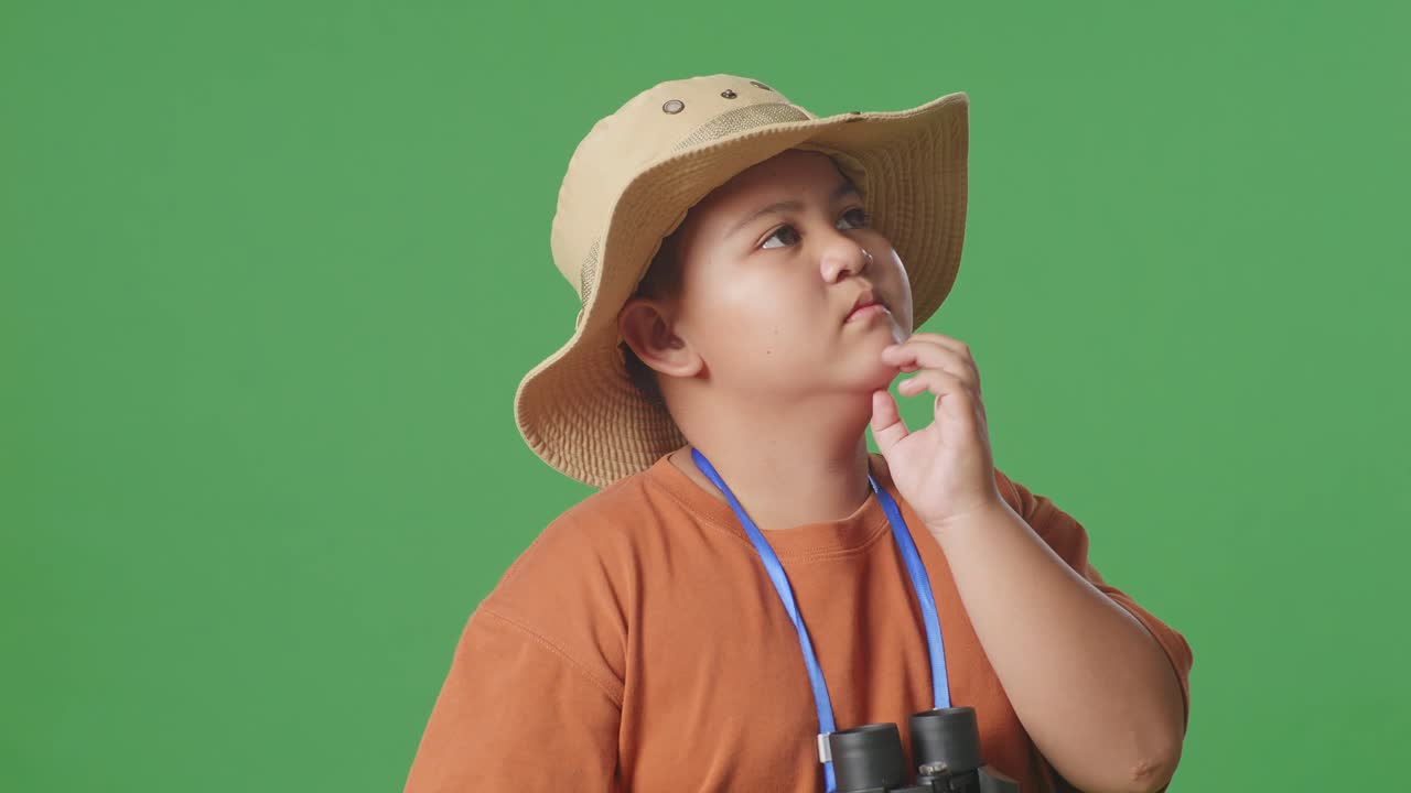 모자와 안경을 입은 아시아 관광 소년은 녹색 스크린 배경에 서서 무언가를 생각하고 주변을 둘러보고 있습니다. 소년 연구자, 여행 관광 모험 개념, 클로즈업
