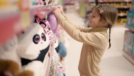 the girl is standing in a children's toy store in front the shelf and take a soft toy