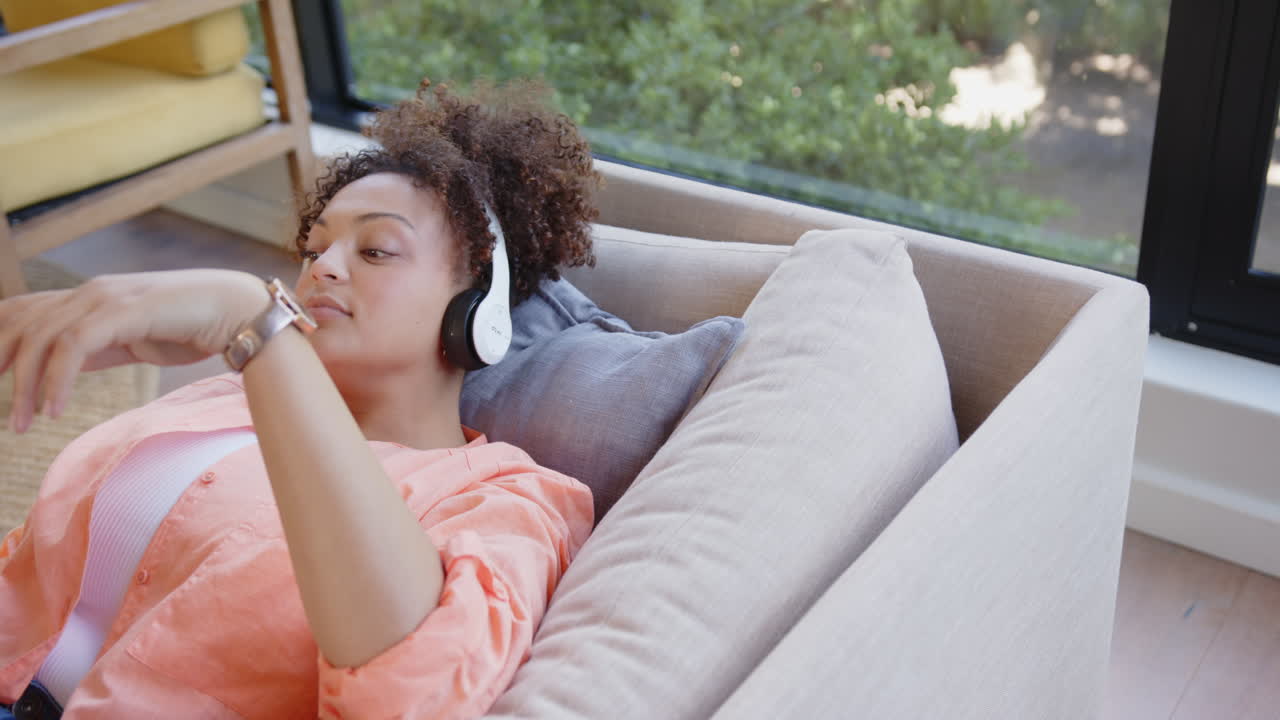 Wearing headphones and using smartphone, woman relaxing on couch enjoying leisure time