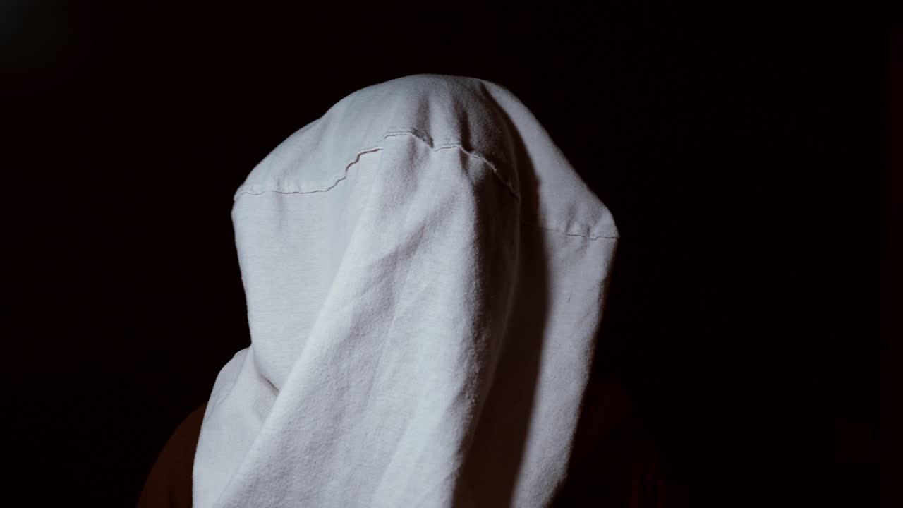 A person sits in darkness with a white cloth covering their head, illuminated by a focused white light. The scene creates a tense and cinematic atmosphere suitable for crime