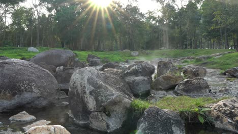 4k static shot of natural hot springs flow along the rocks under sunlight and sun flares, the steam floating up with tree and grass background. chaeson national park, lampang,thailand.