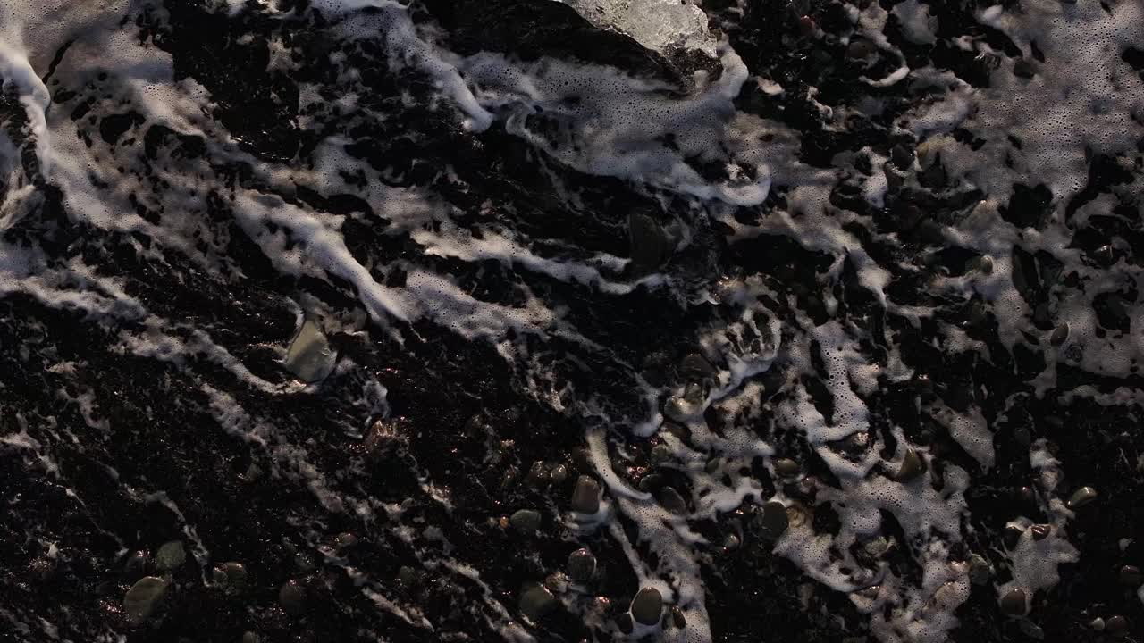 Foamy waves crash over black pebbles and melting ice on Iceland’s Diamond Beach