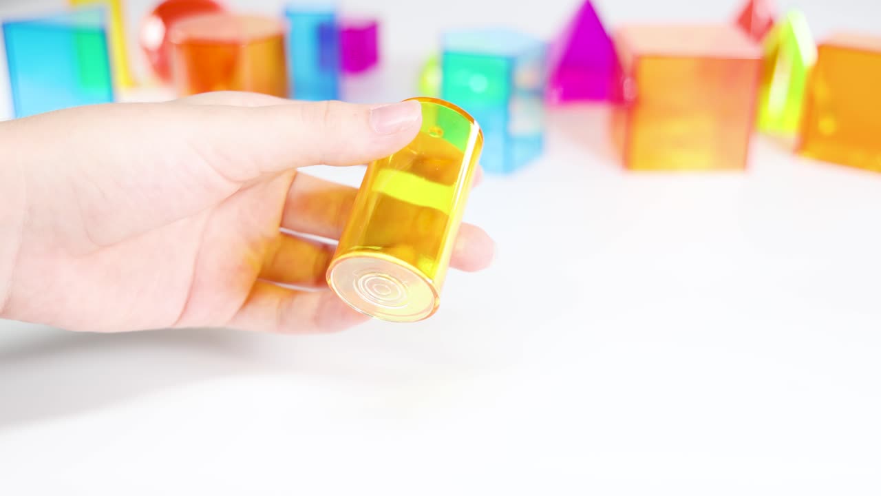 Hands interact with vibrant 3D geometric shapes on a white table, emphasizing educational play and tactile learning