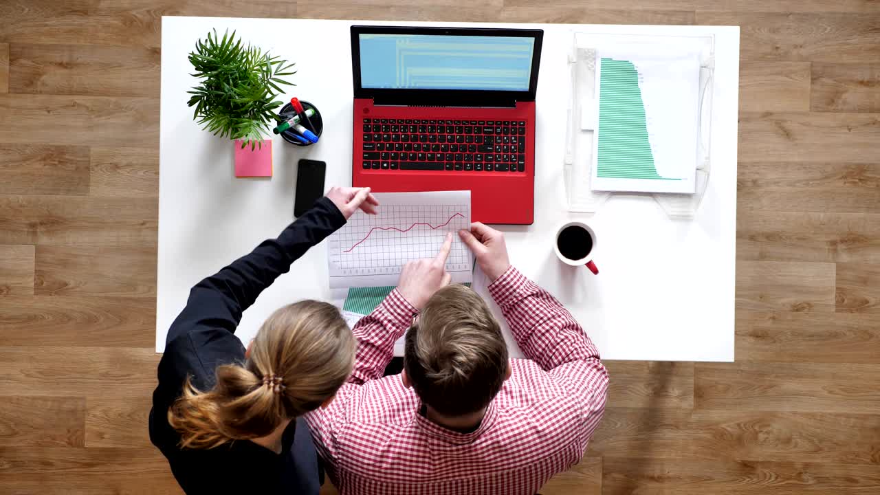 joven con gafas y mujer hablando y señalando en el documento, topshot, escritorio con café y portátil