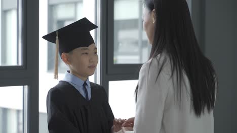 asian kid holding hands with his mother. they hug. he is wearing a gown and a mortarboard.