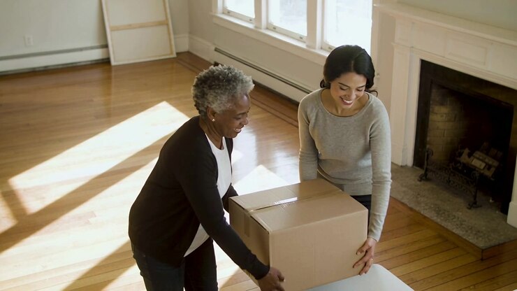 Carrying cardboard box two women in cardigan and jeans placing on living room table preparing move