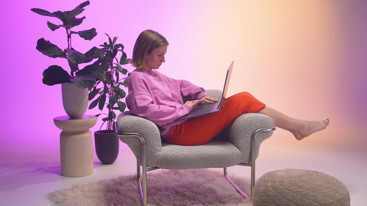 A cheerful woman types on her laptop while reclining barefoot in a cozy chair. Dressed in pink and orange, she enjoys a calm, colorful work moment in a modern space filled with soft textures