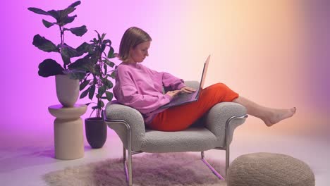 a cheerful woman types on her laptop while reclining barefoot in a cozy chair. dressed in pink and orange, she enjoys a calm, colorful work moment in a modern space filled with soft textures
