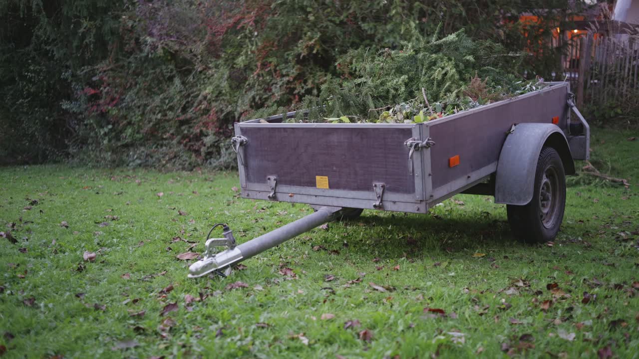 Vista frontale diagonale di un rimorchio in un giardino tradizionale tedesco, che mostra completamente il timone ed è pieno di talee di alberi e arbusti, evidenziando la manutenzione stagionale del giardino e le trame esterne