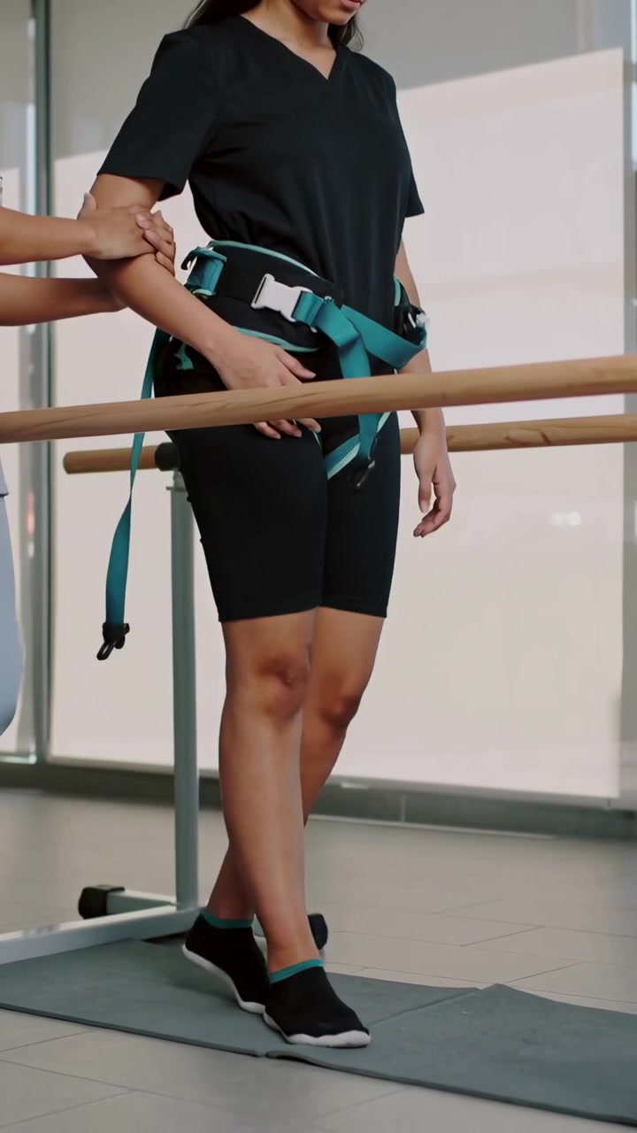 Vertical video: Therapist steadying patient in teal harness stepping along parallel bars in clinic