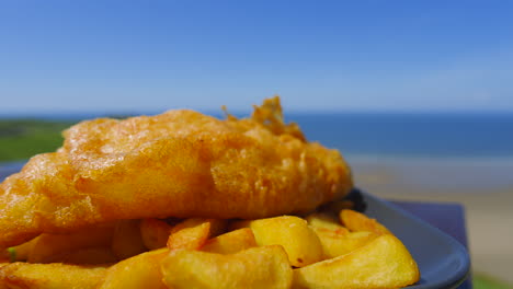 battered fish resting on chunky chips ready to be eaten on british seaside with beautiful blue sea and sky. classic english fast food cuisine.
