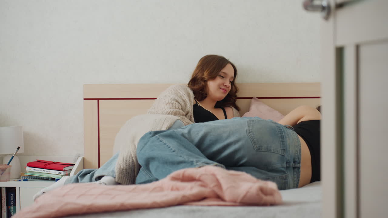 Young Woman In Cozy Attire Exploring Phone, Young Woman Resting Comfortably While Casually Scrolling Her Device, Youthful Woman Dressed In Casual Clothes Enjoying Quiet Moment With Her Phone In Bed