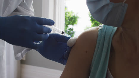 close up of male african american doctor wearing face mask injecting covid-19 vaccine into senior fe