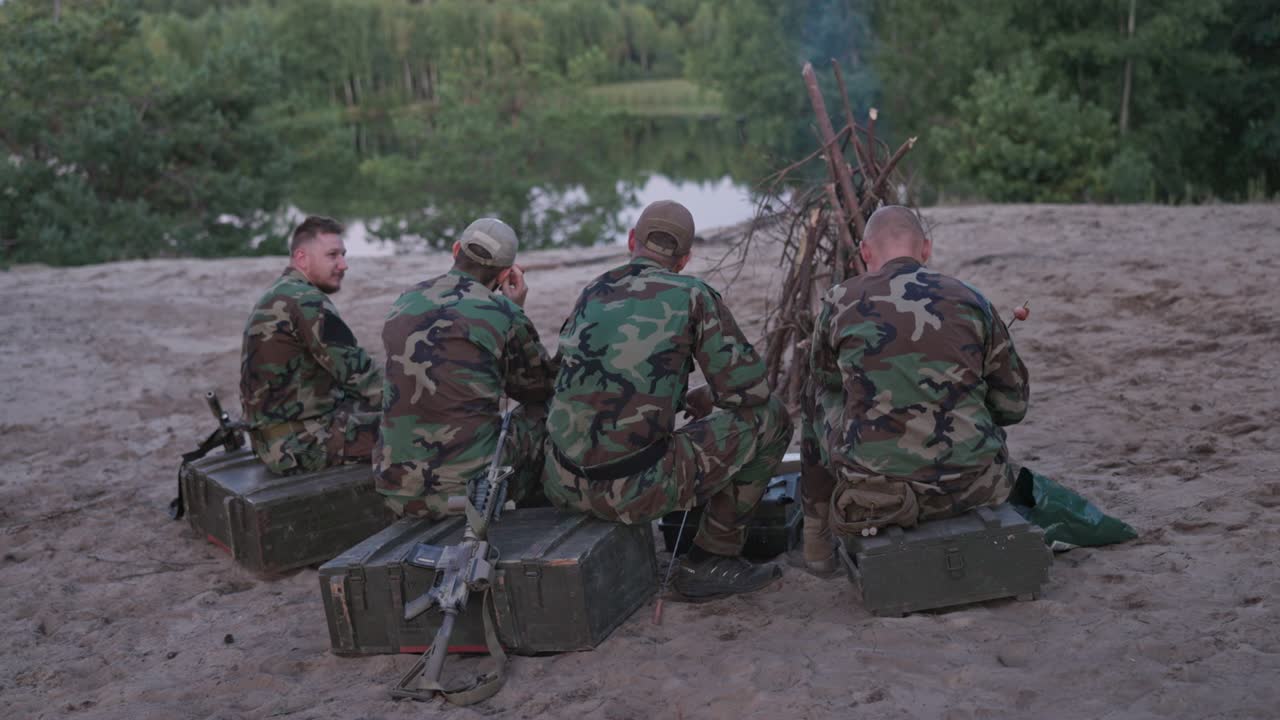 cazadores vestidos con ropa de camuflaje descansan junto al lago, celebrando la caza de caza, se enciende una hoguera, los hombres están fritando salchichas en palos, detrás de ellos las armas se apoyan en cajas