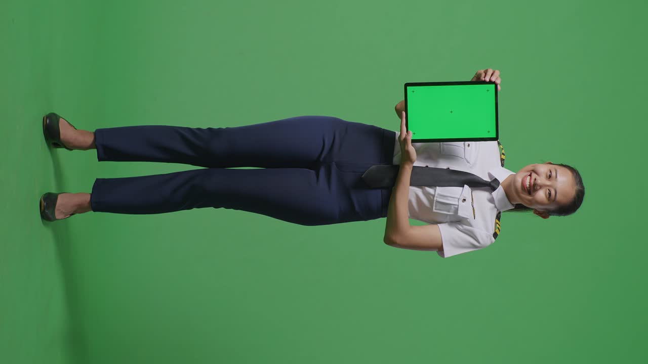 cuerpo lleno de piloto mujer asiática sonriendo y mostrando la tableta de pantalla verde a la cámara mientras estaba de pie en el estudio de fondo de pantalla verde