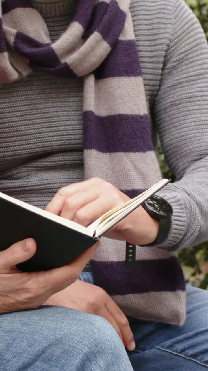 Video verticale: Uomo in maglione che apre un quaderno, indicando le pagine e leggendo su una panchina